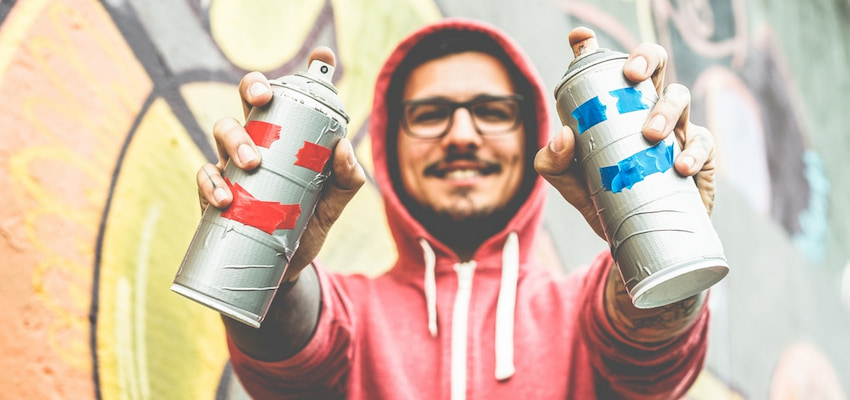 employee holds up two spray cans in front of a graffiti wall