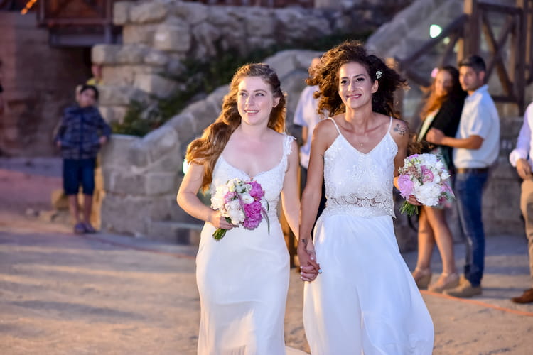 Two brides carrying flowers as they walk down aisle outside