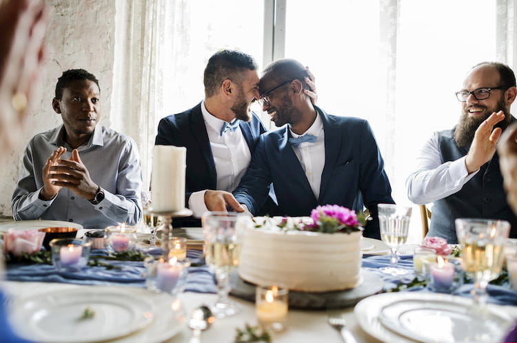 Two grooms kissing at table with wedding cake and guests