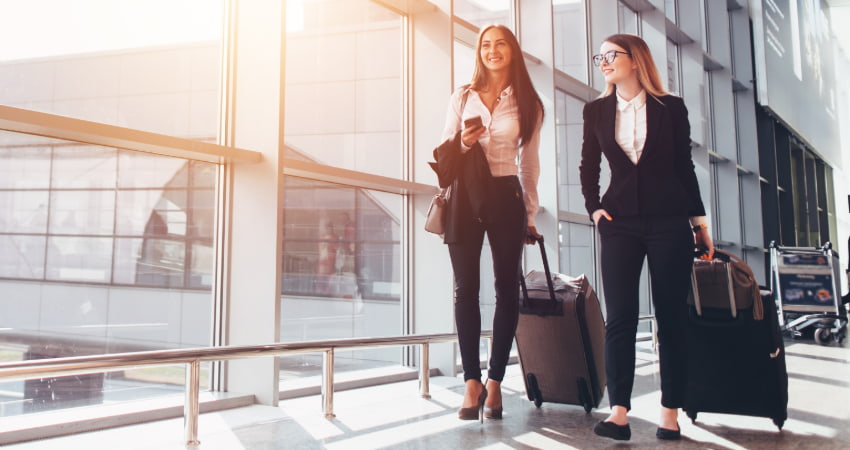 two women walk through an airport with suitcases and suit jackets