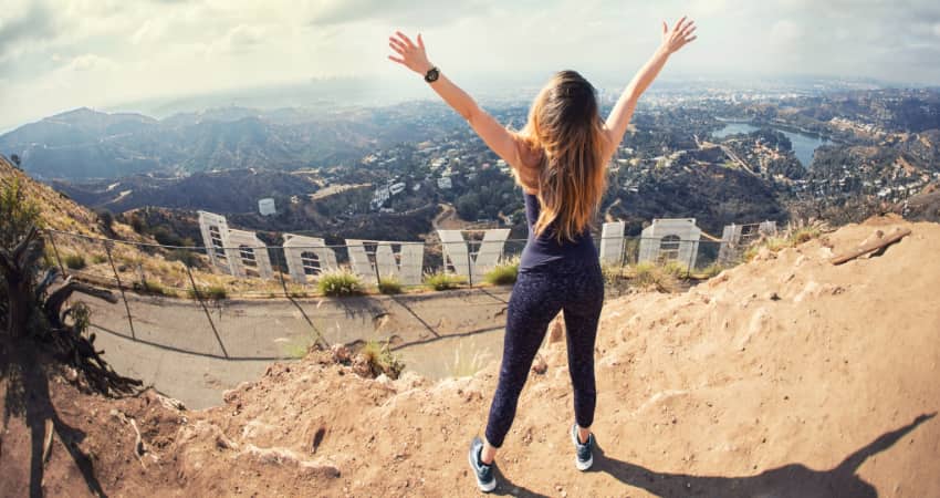 A woman stands on the peak behind the Hollywood sign with arms outstretched