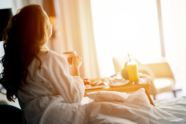 A girl sitting on a hotel bed sipping a drink and staring at the early morning sunshine