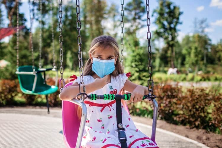 A young girl is wearing a blue face mask while sitting on a swing