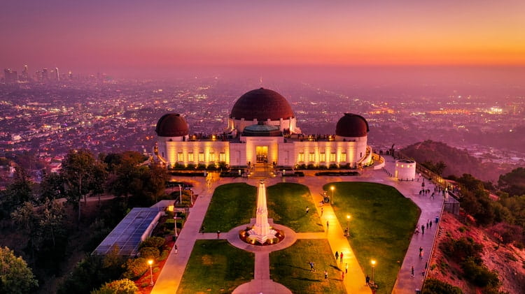 an aerial view of the observatory's front lawn complex and building, at dusk, with los angeles in the background