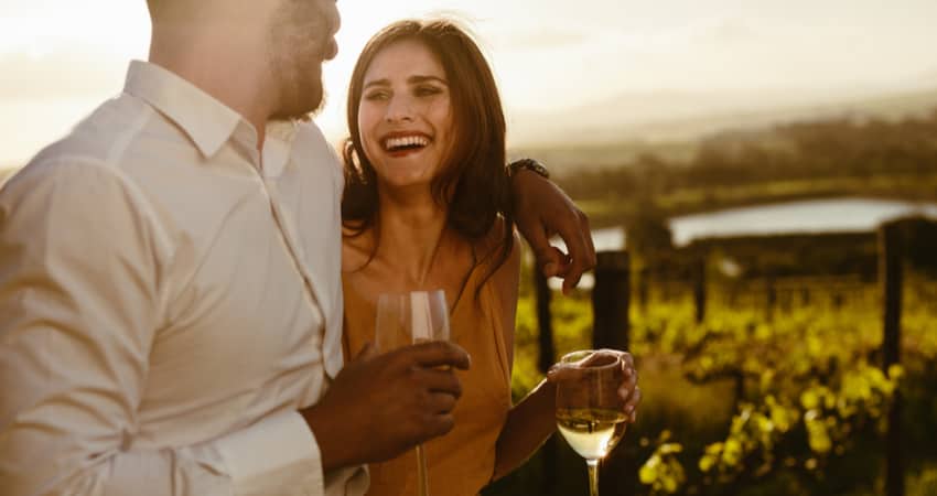 A couple smiling and drinking white wine in front of a vineyard