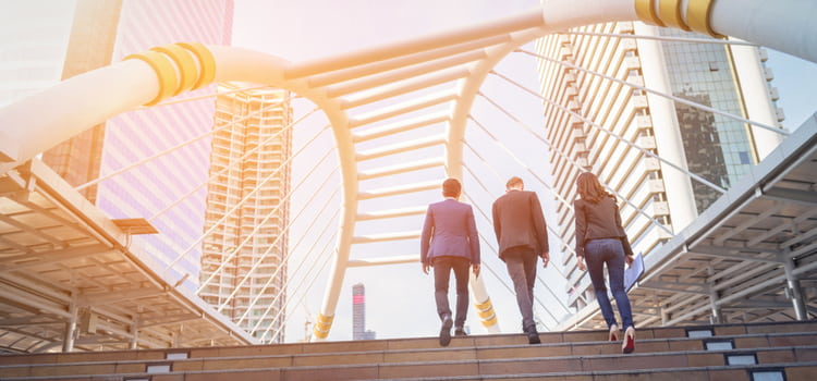 three businesspeople walk up to a large bridge structure with the sun shining low in the background