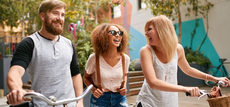 three friends with bikes smile while they walk through a shaded city street