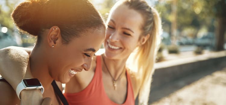 two friends smile in a park near a fountain while one puts their arm around the other