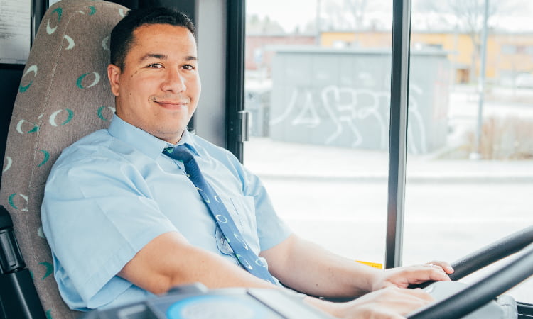 A bus driver sits in the driver seat, hands on the wheel and smiling